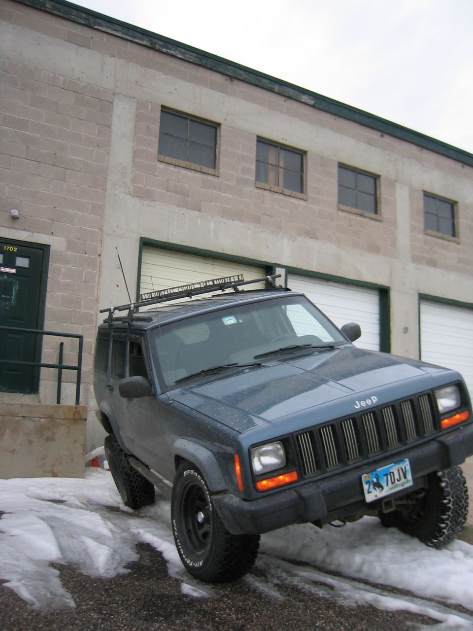 Lifted blue-gray Jeep Cherokee XJ on snowy lot with roof rack, light bar and all-terrain tires