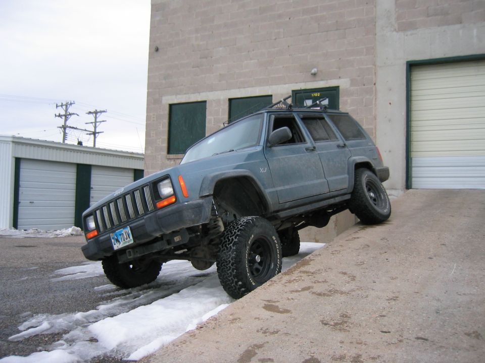 Lifted blue-gray Jeep Cherokee XJ flexing on a concrete ramp with mud tires and roof rack