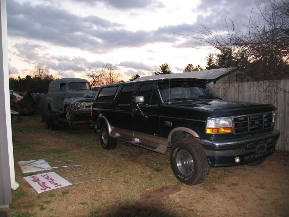 Dark Ford crew cab pickup with topper beside vintage gray panel van on trailer at dusk