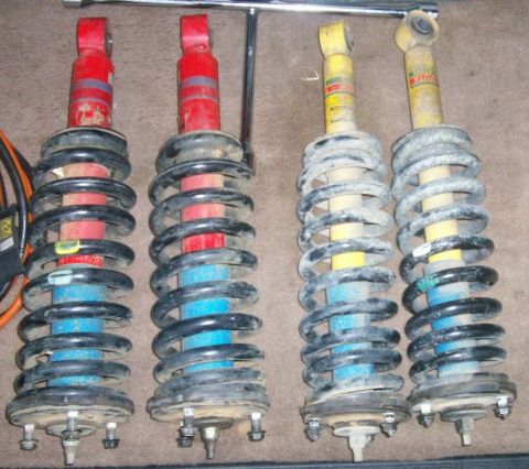 1993 Ford Bronco - Four removed coil-over shock and spring assemblies laid on carpet for comparison: a pair with red-bodied shocks and black springs on the le…