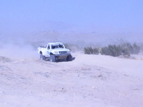 White off-road race truck driving over a sand berm in dusty desert