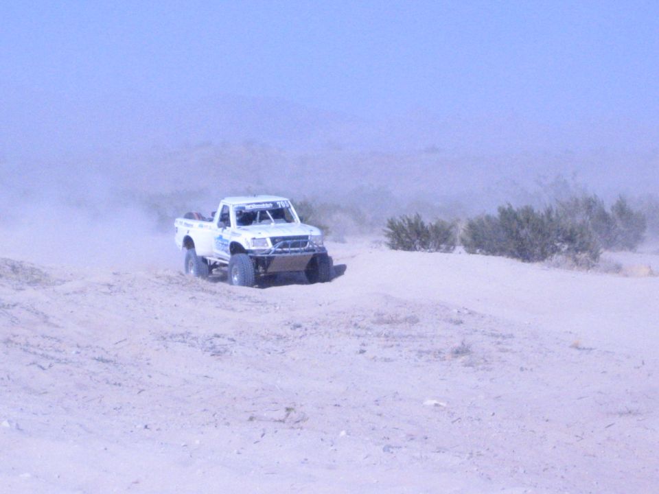 White off-road race truck driving over a sand berm in dusty desert