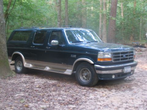 1995 Ford C-150 (Centurion Conversions) - A dark green Ford Bronco SUV with tan trim is parked on a dirt path in a wooded area.
