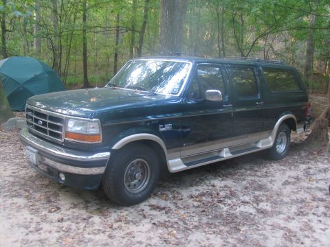 1995 Ford C-150 (Centurion Conversions) - A dark green 1995 Ford Bronco with tan trim is parked in a wooded area near a tent.