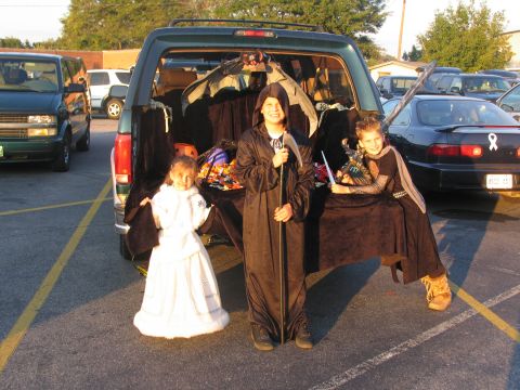 1995 Ford C-150 (Centurion Conversions) - Three children in costumes stand near an open SUV, ready for a trunk-or-treat event.