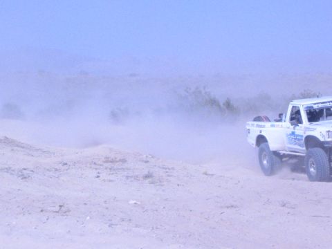 White off-road race truck at right amid heavy dust on sand course