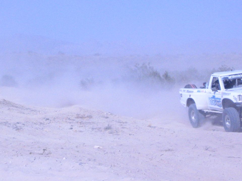 White off-road race truck at right amid heavy dust on sand course