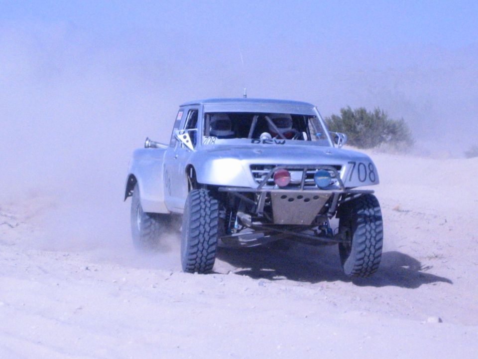 Silver number-708 off-road race truck head-on kicking up sand