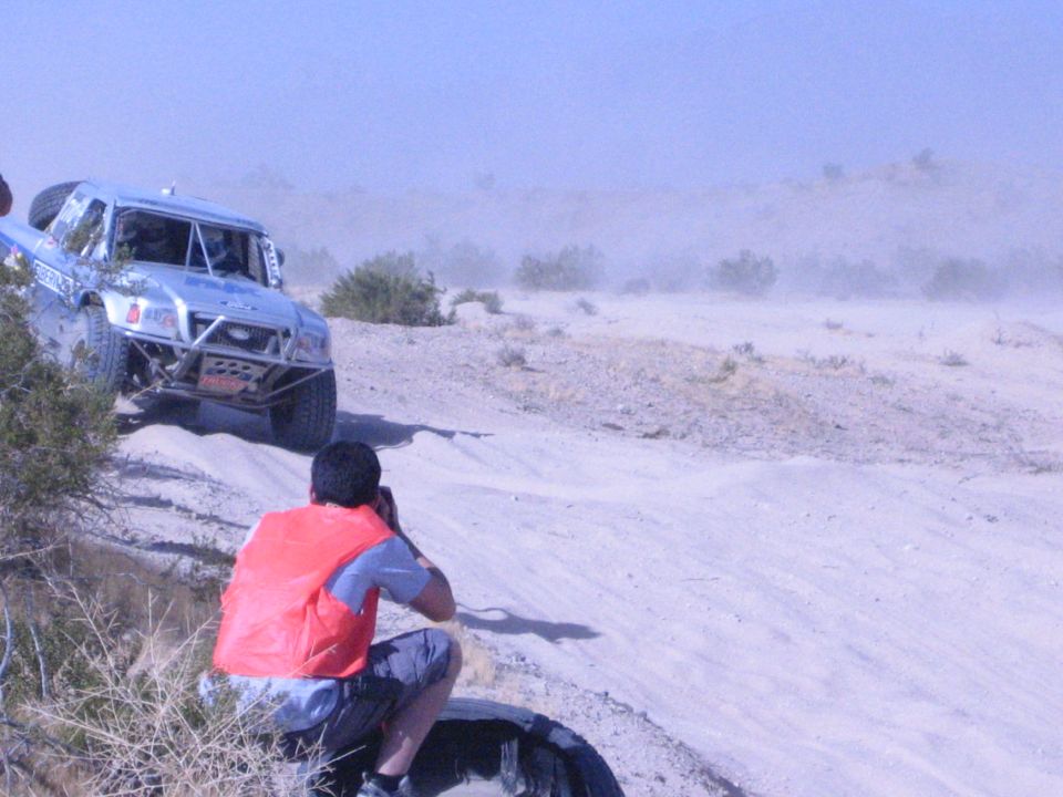 Photographer in orange vest shooting a silver race truck in the desert