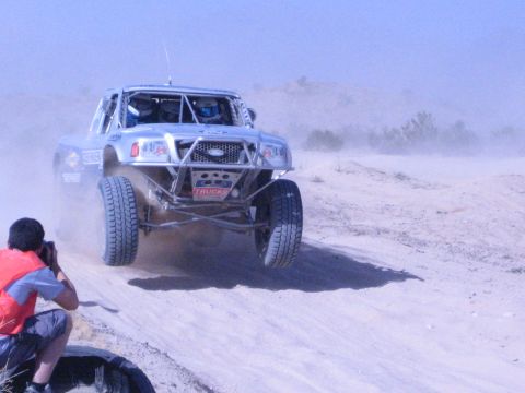 Silver off-road race truck launches over a sandy berm kicking up dust in the desert