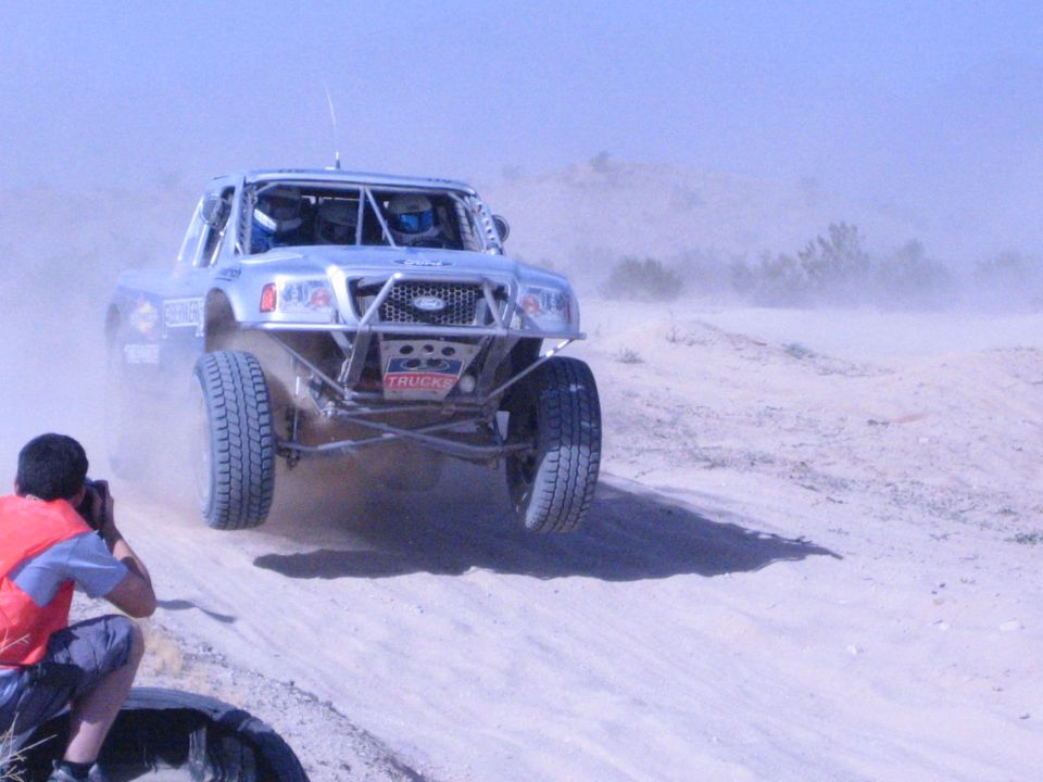 Silver off-road race truck launches over a sandy berm kicking up dust in the desert