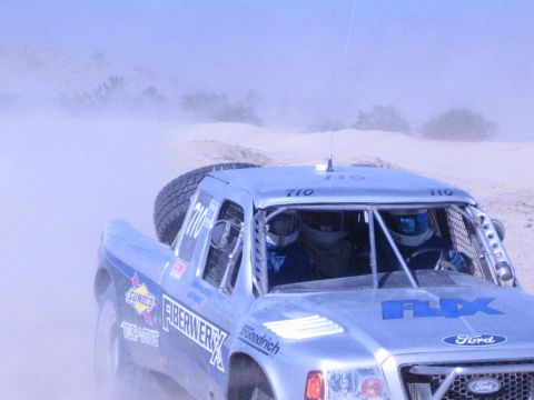 Close view of silver Ford race truck 710 cockpit with two helmeted occupants in dust