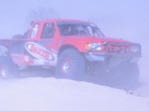 Red off-road race truck with Maxxis graphics on a sandy crest in heavy dust haze