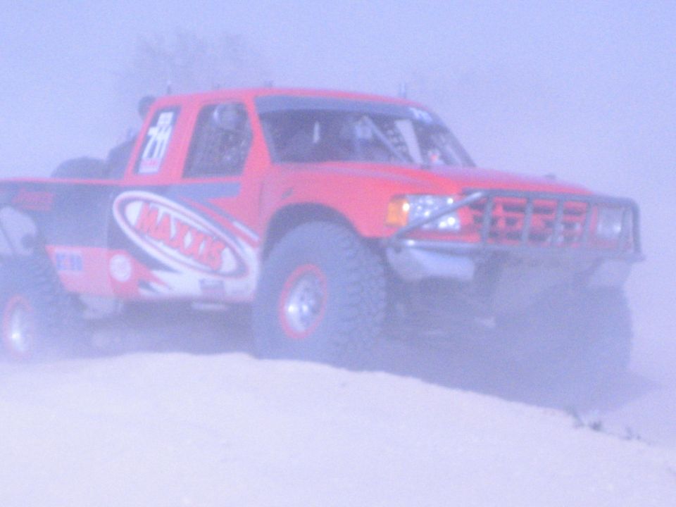 Red off-road race truck with Maxxis graphics on a sandy crest in heavy dust haze