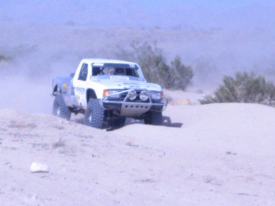 White off-road race truck climbing a sandy desert berm amid dust and brush