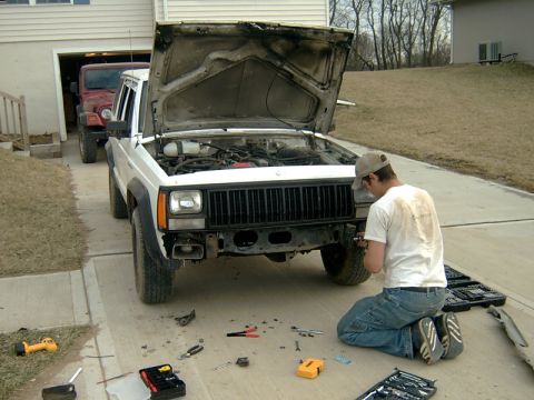 1998 Jeep TJ - A man works on the front of a white Jeep Cherokee XJ while a red Jeep Wrangler is parked behind it.