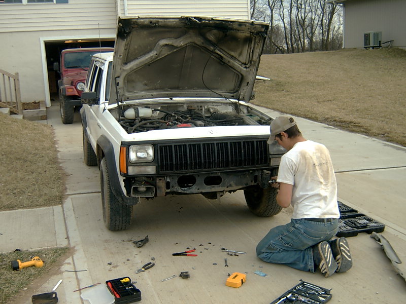 White Jeep Cherokee XJ with hood open, man working on front bumper, tools scattered on driveway. Red Jeep Wrangler in background