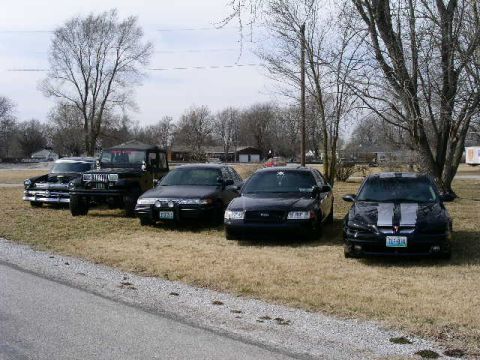 2001 Ford Crown Victoria - A collection of vehicles, including a vintage car, a Jeep, and two modern sedans, are parked outdoors on a dry, grassy area.