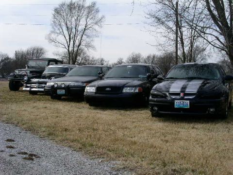 2001 Ford Crown Victoria - Several vehicles are lined up in a grassy area with trees in the background.