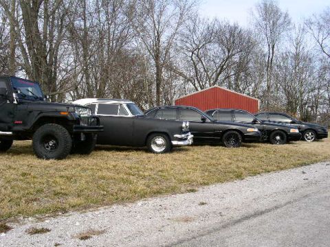 2001 Ford Crown Victoria - A collection of black vehicles, including a Jeep and several sedans, are lined up outdoors near a red barn.
