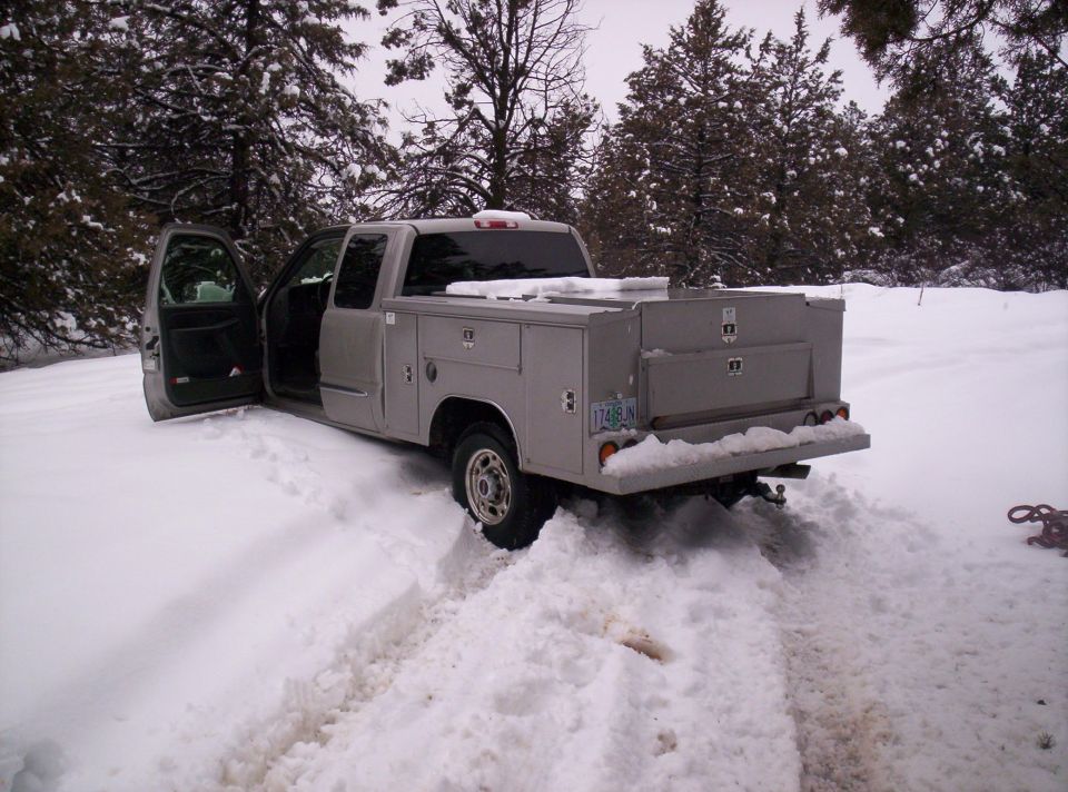 Silver utility-bed pickup truck stuck in deep snow with the driver door open