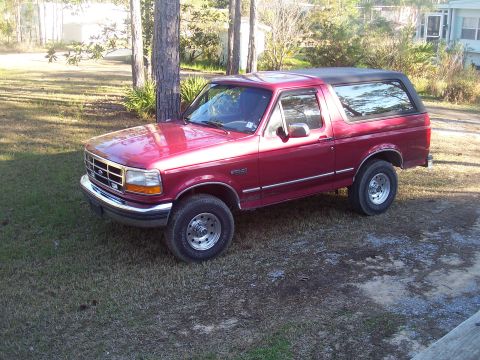 1995 Ford Bronco - Driver-side three-quarter front view of a red Ford Bronco with a black hardtop, parked on a grassy pine lot beside a light-colored house. C…
