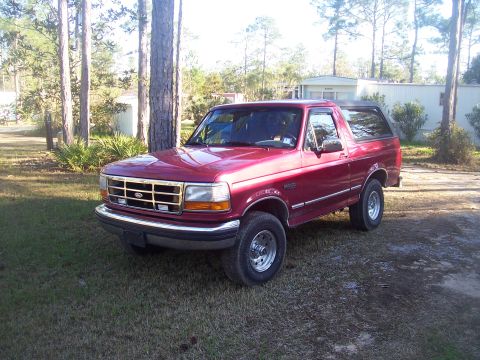 1995 Ford Bronco - Three-quarter front view of a red Ford Bronco with a black hardtop parked on a grassy pine-shaded lot next to a white mobile home. It shows…