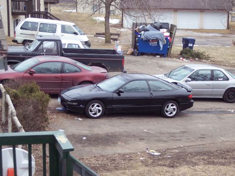 1995 Ford Probe - Several cars are parked in a lot with a dumpster overflowing with trash in the background.