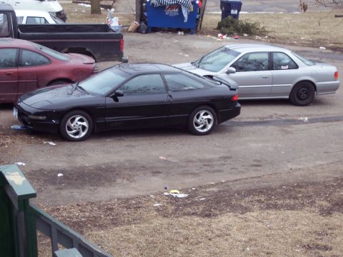 1995 Ford Probe - A black coupe with distinctive pop-up headlights is parked on a dirt lot next to other vehicles.