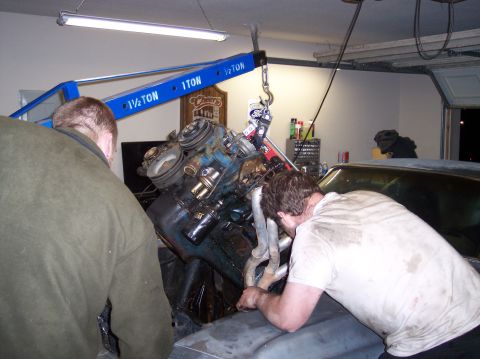1967 Ford Mustang - Two mechanics work on an engine being hoisted out of a vehicle in a garage setting.