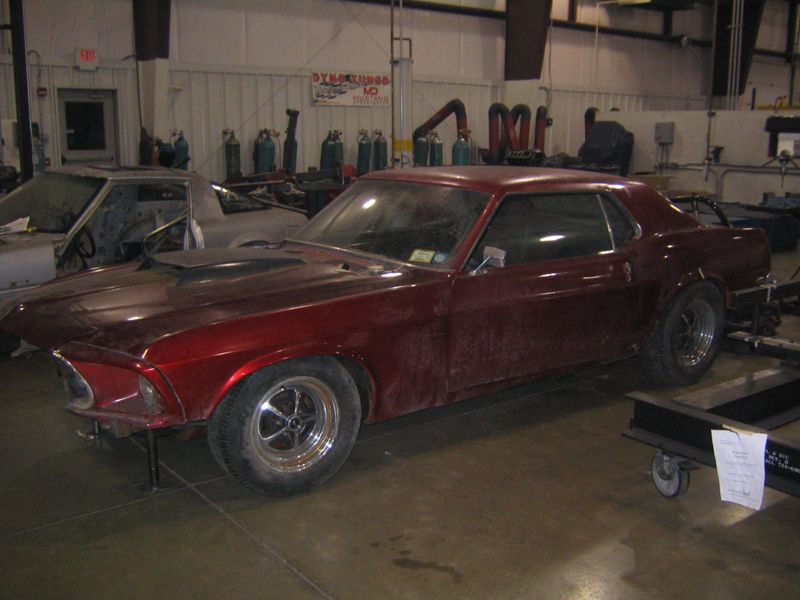 Red 1969 Ford Mustang coupe with a scoop hood, in a workshop. Front bumper is missing
