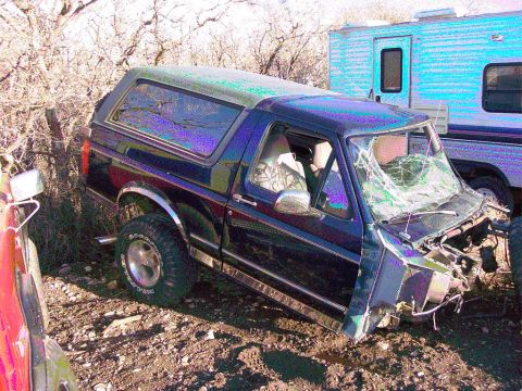 1981 Ford F-150 - A damaged dark blue Ford Bronco sits in a dirt lot next to a trailer.