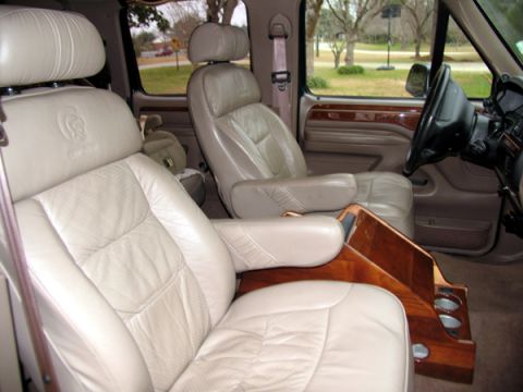 1995 Ford C-150 (Centurion Conversions) - The interior of a van shows plush tan leather captain's chairs and a wooden center console with cup holders.