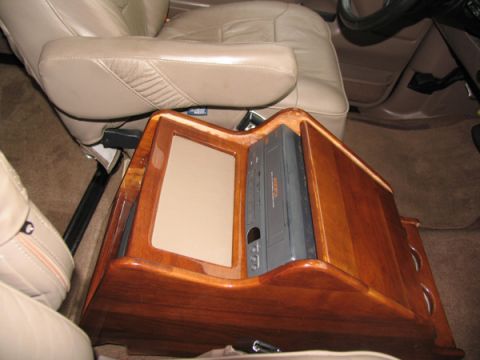 1995 Ford C-150 (Centurion Conversions) - The interior of a vehicle shows a custom wooden console with a VCR and a screen, situated between tan leather seats.