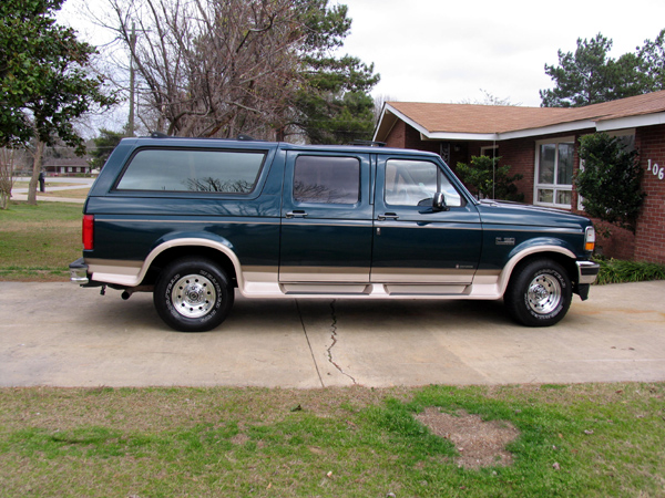 Green and tan Ford conversion SUV parked in profile on a driveway by a brick house.