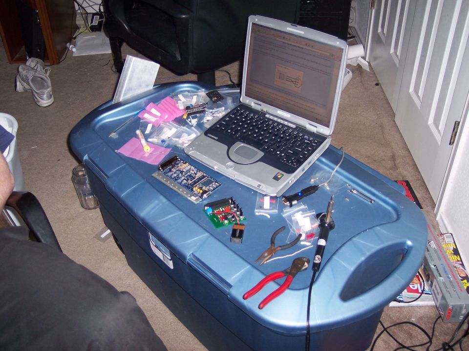 Laptop open on a blue plastic bin with electronic components, tools, and bags of parts scattered around
