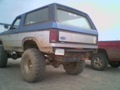 1985 Ford Bronco - A lifted Ford Bronco with large tires is parked next to a red truck.