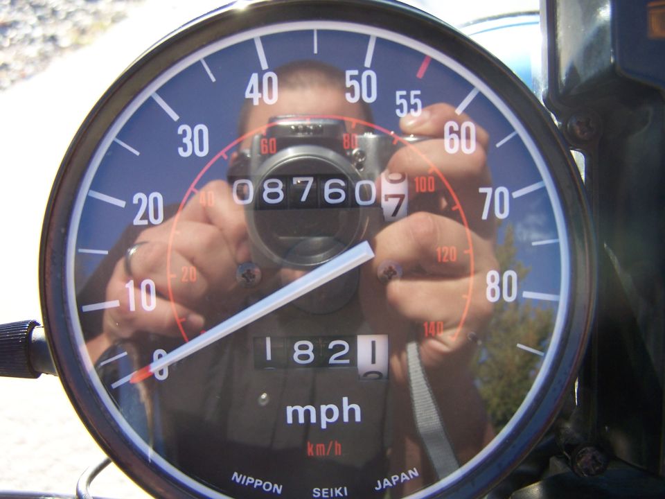 Motorcycle speedometer with odometer reading 1821, reflecting a person holding a camera