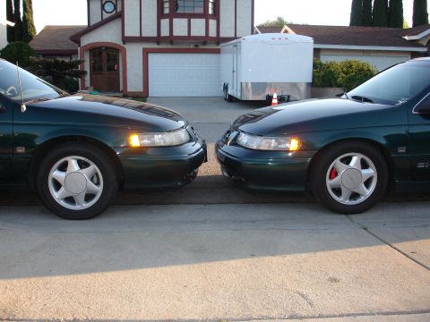 1995 Ford Taurus - Two dark green 1995 Ford Tauruses are parked facing each other on a suburban street in front of a house.