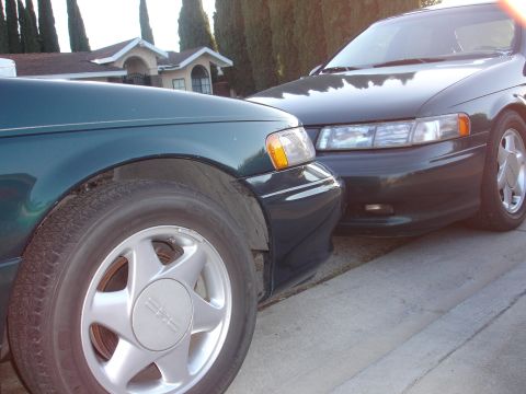 1995 Ford Taurus - Two dark sedans are parked side-by-side on a residential street.