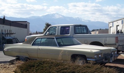 1972 Ford Ranchero - A 1972 Cadillac Coupe DeVille is parked next to a pickup truck with a mountain range in the distance.