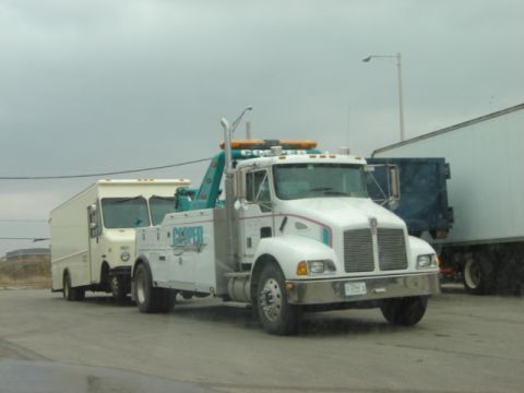 1992 Ford Bronco - A white tow truck is hauling a white box truck on a cloudy day. A large trailer is visible in the background.
