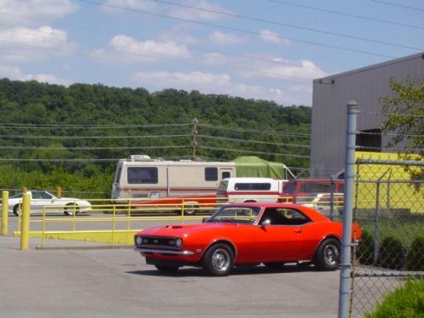 1979 Ford Bronco - A red 1967 Chevrolet Camaro coupe is parked in front of a building with RVs and a white car in the background.