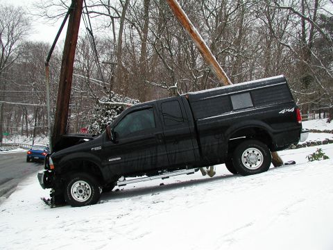 1992 Ford Taurus - Black Ford F-250 Super Duty pickup truck crashed into a utility pole in snowy conditions. Another vehicle is on the road.