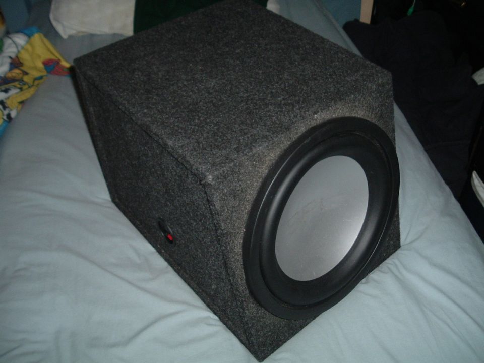 Carpeted wedge subwoofer enclosure with silver-cone driver and side terminal cup on a bed