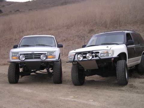 1985 Ford Bronco II - Two modified Ford Explorer SUVs with off-road bumpers and large tires are parked on a dirt road.