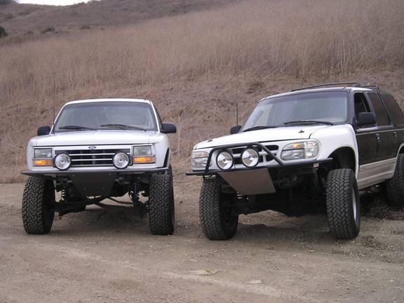 Two modified Ford Explorer SUVs parked on a dirt road, one white, one white and black