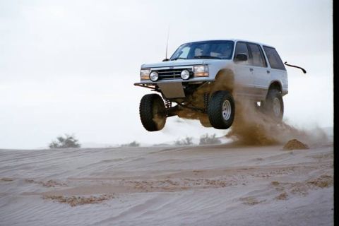 1985 Ford Bronco II - A modified SUV soars through the air above sandy terrain, creating a cloud of dust.
