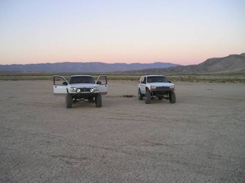 1985 Ford Bronco II - Two SUVs are parked in a vast, dry, cracked earth landscape with mountains in the background at sunset.