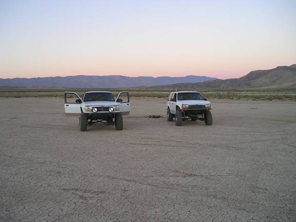 Two white SUVs parked in a dry lakebed at dusk. The SUV on the left has its doors open